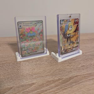 May include: Two Pokemon trading cards in clear plastic holders, displayed on white stands. The cards feature colorful illustrations of Venusaur EX and Zeraora VMAX. The Venusaur card shows a green and pink creature, while the Zeraora card is yellow and blue.