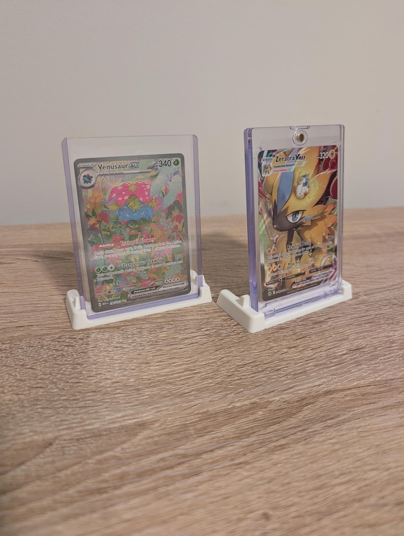 May include: Two Pokemon trading cards in clear plastic holders, displayed on white stands. The cards feature colorful illustrations of Venusaur EX and Zeraora VMAX. The Venusaur card shows a green and pink creature, while the Zeraora card is yellow and blue.