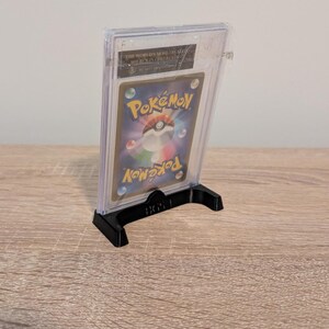 May include: A graded Pok&eacute;mon trading card in a clear plastic case, displayed on a black stand. The card features the Pok&eacute;mon logo and a colorful design. The stand is labeled "BGS 1".