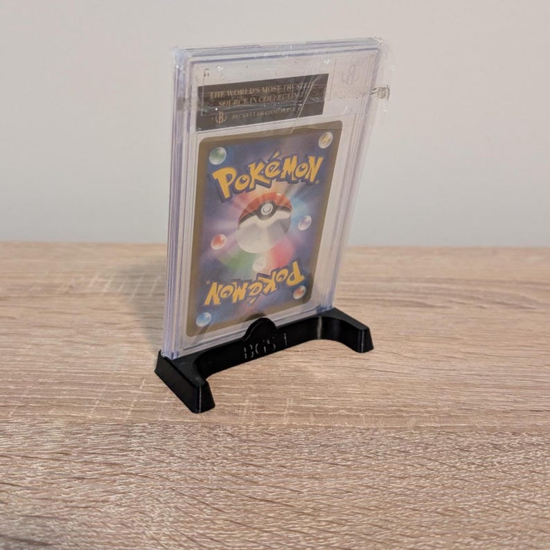 May include: A graded Pok&eacute;mon trading card in a clear plastic case, displayed on a black stand. The card features the Pok&eacute;mon logo and a colorful design. The stand is labeled "BGS 1".