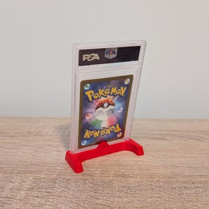 May include: A graded Pok&eacute;mon trading card in a clear plastic case, displayed on a red stand. The card features the Pok&eacute;mon logo and a colorful design. The case has the PSA logo.