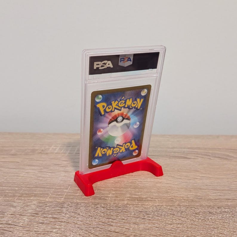 May include: A graded Pok&eacute;mon trading card in a clear plastic case, displayed on a red stand. The card features the Pok&eacute;mon logo and a colorful design. The case has the PSA logo.