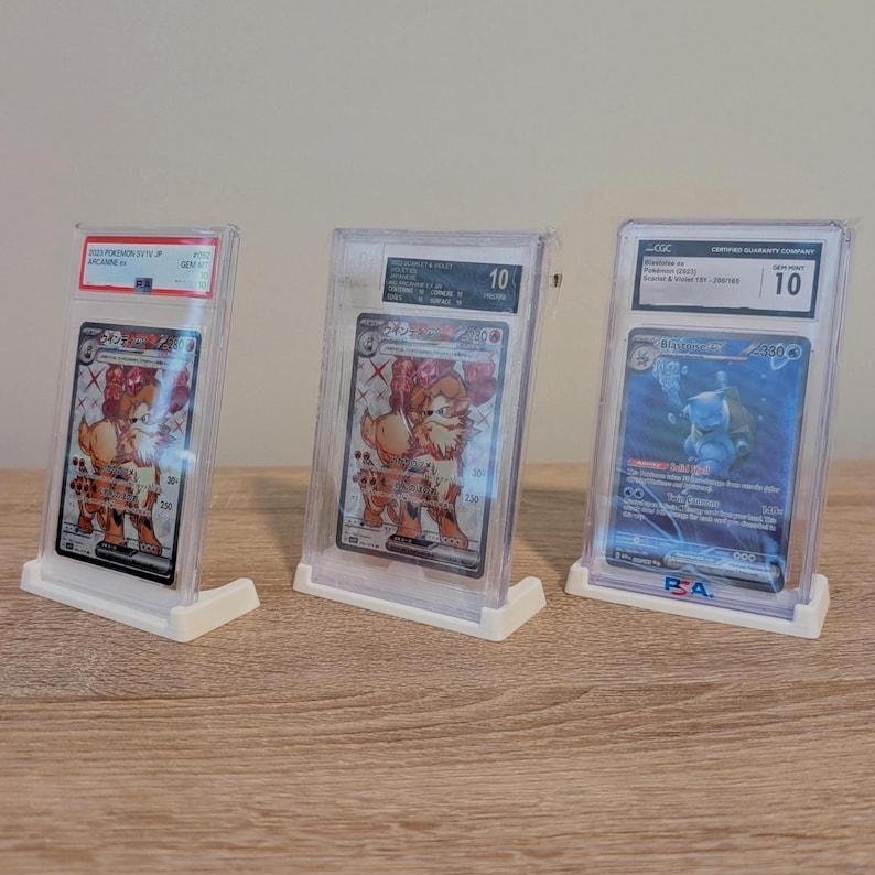 May include: Three graded Pok&eacute;mon trading cards in clear plastic cases, displayed on white stands. The cards feature colorful illustrations of Pok&eacute;mon characters. The cases have labels with grades and certification information.