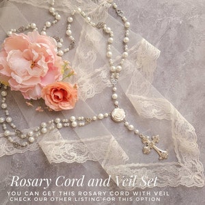 May include: A white pearl rosary with a silver cross and a white rose charm, displayed on a white lace fabric with pink flowers.