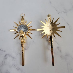 May include: Two gold-colored crucifixes. One features a detailed depiction of Jesus Christ on the cross, surrounded by a sunburst design. The other shows the back of the crucifix with a sunburst design. The cross has the text "DIO E AMORE" and "IHS".