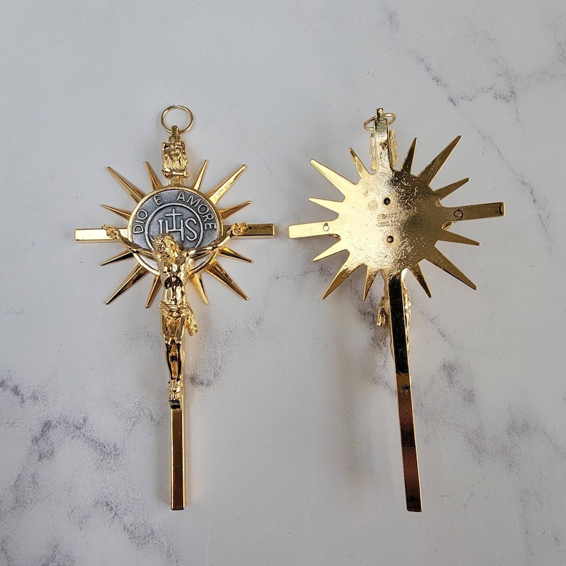 May include: Two gold-colored crucifixes. One features a detailed depiction of Jesus Christ on the cross, surrounded by a sunburst design. The other shows the back of the crucifix with a sunburst design. The cross has the text "DIO E AMORE" and "IHS".