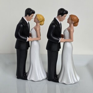 May include: Two wedding cake toppers. Each features a groom in a black tuxedo and a bride in a white wedding dress. The figures are facing each other, holding hands, and looking into each other's eyes. One bride has blonde hair, the other has red hair.
