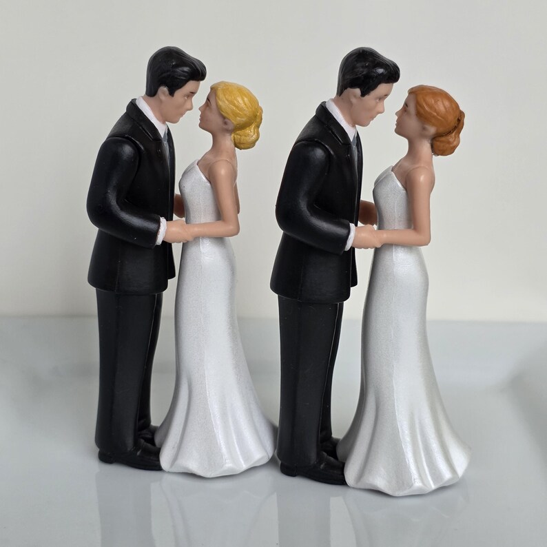 May include: Two wedding cake toppers. Each features a groom in a black tuxedo and a bride in a white wedding dress. The figures are facing each other, holding hands, and looking into each other's eyes. One bride has blonde hair, the other has red hair.