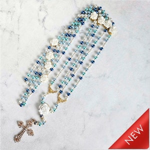 May include: Several rosaries with white rose accents and a cross pendant. The rosaries feature a mix of blue, white, and pearl-colored beads. The cross pendant is embellished with rhinestones. The background is a light, textured surface.