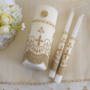 May include: Three white wedding candles with gold lace trim and a pearl embellishment. The center candle has a gold ring charm.