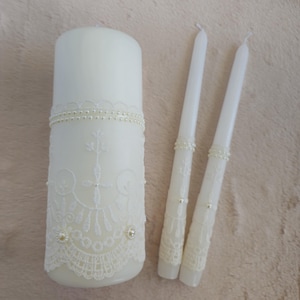 May include: Three white wedding unity candles with lace and pearl accents. The larger candle is taller and has a wider base. The two smaller candles are thinner and shorter.