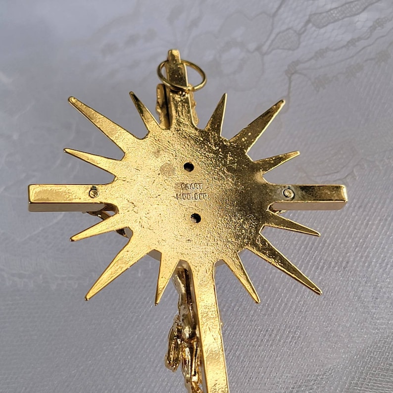 May include: A gold-toned crucifix pendant with a sunburst design. The back of the cross features the text "CAART - MOLDED". The crucifix depicts a detailed figure of Jesus Christ.