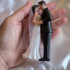 May include: A wedding cake topper featuring a bride and groom figurine. The bride wears a white strapless gown and veil, while the groom is in a black tuxedo. The figurines are kissing, holding hands, and are approximately 4 inches tall.