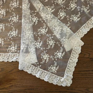 Può includere: Un tessuto in pizzo trasparente color bianco sporco caratterizzato da un intricato motivo floreale ricamato. I bordi sono rifiniti con un'ampia bordura in pizzo smerlato e un piccolo fiocco in nastro bianco è fissato a un angolo della decorazione.
