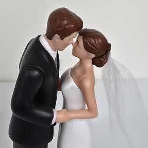 May include: A wedding cake topper featuring a bride and groom figurine. The groom is in a black tuxedo, and the bride wears a white, one-shoulder gown and a veil. Both figures have brown hair and are posed close together, touching noses.