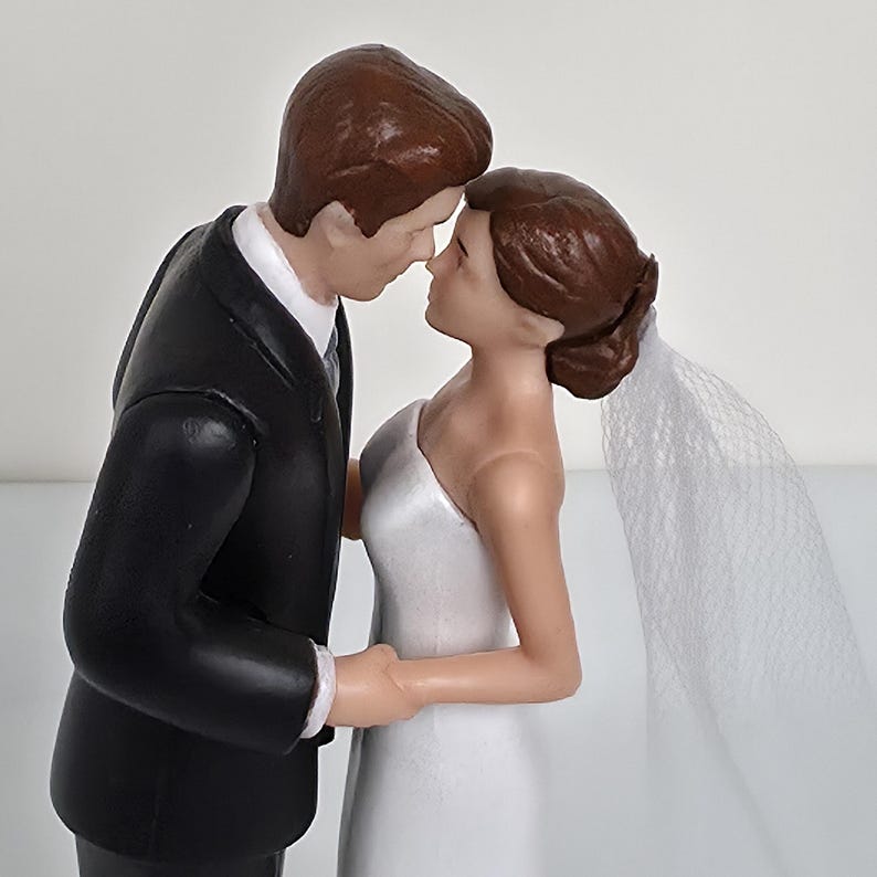 May include: A wedding cake topper featuring a bride and groom figurine. The groom is in a black tuxedo, and the bride wears a white, one-shoulder gown and a veil. Both figures have brown hair and are posed close together, touching noses.