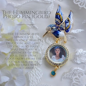 May include: A gold photo pin featuring a blue and white hummingbird with rhinestones. The pin has a round locket with a clear glass window to insert a photo. The locket is attached to the hummingbird by a gold chain. The pin has a small blue gemstone dangling from the bottom. The text "THE HUMMINGBIRD PHOTO PIN (GOLD)" is at the top of the image. The text "THE HUMMINGBIRD SYMBOLIZES INTELLIGENCE, BEAUTY, DEVOTION, AND LOVE. LEGEND ALSO HAS IT THAT THEY ARE A MESSENGER OF THE SOUL, THEY BRING GOOD NEWS FROM THE OTHER SIDE, FROM LOVED ONES WHO PASSED AWAY." is at the bottom of the image.