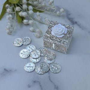 Può includere: Una scatola in filigrana d'argento con una rosa bianca sopra, circondata da piccole monete d'argento con incisioni dettagliate. Un rametto di fiori bianchi con foglie verdi è sullo sfondo. La scatola è rettangolare e ha una piccola chiusura.