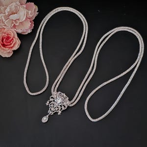 May include: A silver necklace with a large, ornate pendant featuring a central clear stone and a dangling teardrop-shaped stone. The necklace has multiple strands and is displayed on a black surface. Pink flowers are in the background.