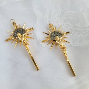 May include: Two gold-colored crucifixes with a detailed depiction of Jesus Christ. Each crucifix features a sunburst design behind the figure and a circular emblem with text. The background is a white, textured fabric.