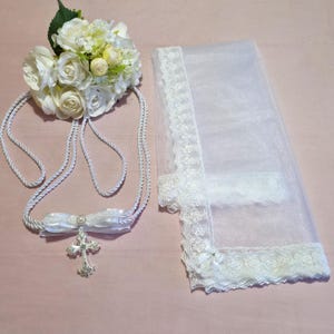 Puede incluir: Velo de encaje blanco con una cinta de satén blanco y un colgante de cruz de plata. El velo está adornado con encaje blanco y tiene un delicado patrón floral. El colgante de cruz está unido a la cinta con una perla.