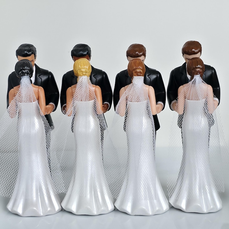 May include: Four wedding cake toppers. Each features a bride in a white gown and veil, standing with a groom in a black tuxedo. The brides have different hair colors: black, blonde, and red. The grooms have dark hair.