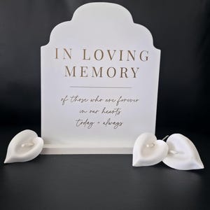 May include: A white memorial sign with the words "IN LOVING MEMORY" and a heartfelt message. Three white heart-shaped calla lilies are at the base. The sign and flowers are set against a dark background.