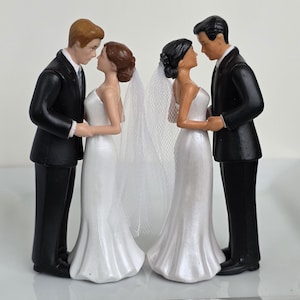 May include: Wedding cake toppers featuring two couples. Each couple is a bride and groom figurine. The brides wear white gowns and veils. The grooms wear black suits. The figurines are holding hands and facing each other.