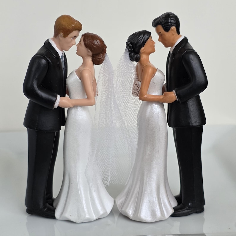 May include: Wedding cake toppers featuring two couples. Each couple is a bride and groom figurine. The brides wear white gowns and veils. The grooms wear black suits. The figurines are holding hands and facing each other.