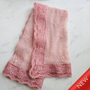 May include: A folded, sheer, blush-pink lace fabric with a darker rose-colored lace border. The fabric has a delicate floral pattern and is labeled "NEW" in the bottom right corner.