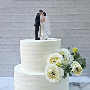 May include: A two-tiered white wedding cake with a textured finish, topped with a figurine of a kissing couple. The cake is decorated with white flowers and green foliage. The background is a white brick wall.