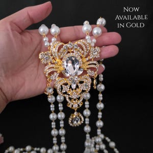 May include: A gold-toned statement necklace with a large, clear gemstone center and smaller clear stones. The necklace features a decorative pendant with intricate detailing and is accented with strands of faux pearls. Text reads "NOW AVAILABLE IN GOLD."