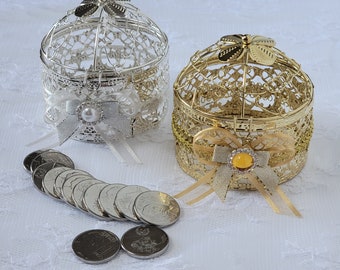 Op de afbeelding: Twee decoratieve, sierlijke vogelkooi-vormige dozen, één zilver en één goud, met lint- en parelaccenten. Een rij zilveren munten is voor de dozen gerangschikt. De dozen staan op een wit, getextureerd oppervlak.