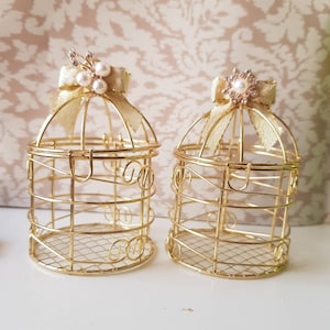 May include: Two decorative gold-colored metal birdcages. Each cage features a bow and a decorative embellishment on top. The cages have a wireframe design with decorative swirls and a small, hinged door. The cages are empty.