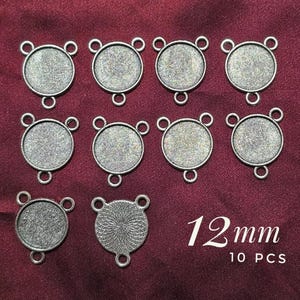 May include: Ten silver-toned metal pendant trays with three loops each. The trays are 12 mm in diameter and have a smooth, slightly textured surface. The text "12 mm 10 PCS" is printed below the trays.