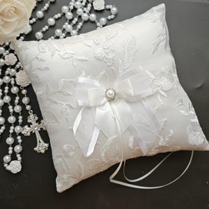May include: White satin ring pillow with a delicate floral embroidery design. A white satin bow with a pearl and rhinestone embellishment is centered on the pillow.