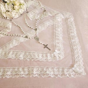 Puede incluir: Velo de encaje blanco con un rosario de perlas y un colgante de cruz de plata. El velo está hecho de encaje delicado y tiene un borde festoneado.