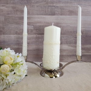 May include: A silver candle holder with three white candles. The center candle is decorated with white lace and pearls. The candles are on a white surface.