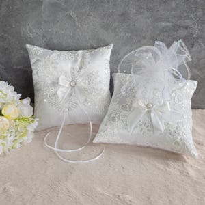 May include: Two white satin ring pillows with lace trim and a pearl button. The pillows are on a white fuzzy surface.