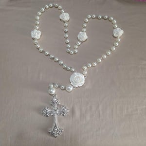 Può includere: Un rosario con perle finte, accenti di rose bianche e un pendente a croce argentata decorativa. La collana è lunga circa 61 cm.