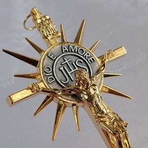 May include: Gold-toned crucifix pendant with a detailed depiction of Jesus Christ on the cross. The pendant features a sunburst design and a central medallion with the inscription "DIO E AMORE" and the letters "IHS".