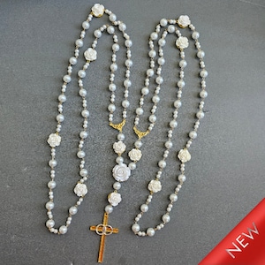 May include: A white pearl rosary necklace with white rose accents and a gold cross pendant.