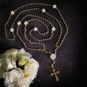 May include: A white pearl rosary with a gold cross and white rose accents. The rosary is laid out on a dark gray background.
