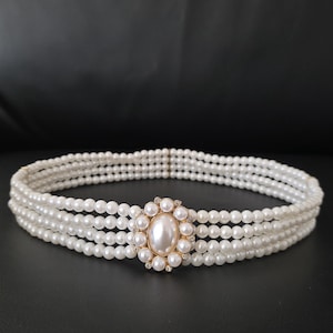 May include: A white pearl choker necklace with four strands of small pearls. The focal point is an oval-shaped gold-toned centerpiece with a large pearl and surrounding smaller pearls and clear crystals.