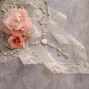 Puede incluir: Un collar de rosario con perlas blancas artificiales, eslabones de metal plateado, un emblema central de rosa blanca y un colgante de cruz plateado ornamentado. El collar est&aacute; sobre una tela de encaje color crema junto a una peon&iacute;a rosa.