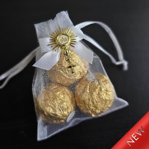 Puede incluir: Bolsa de organza blanca con un amuleto religioso dorado y chocolates envueltos en oro. El amuleto presenta un cáliz y las letras IHS.