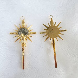 May include: Two gold-colored crucifixes. One shows a detailed depiction of Jesus Christ on the cross, with a sunburst design behind the cross. The other shows the back of the cross with a sunburst design. The crosses have a loop at the top for hanging.
