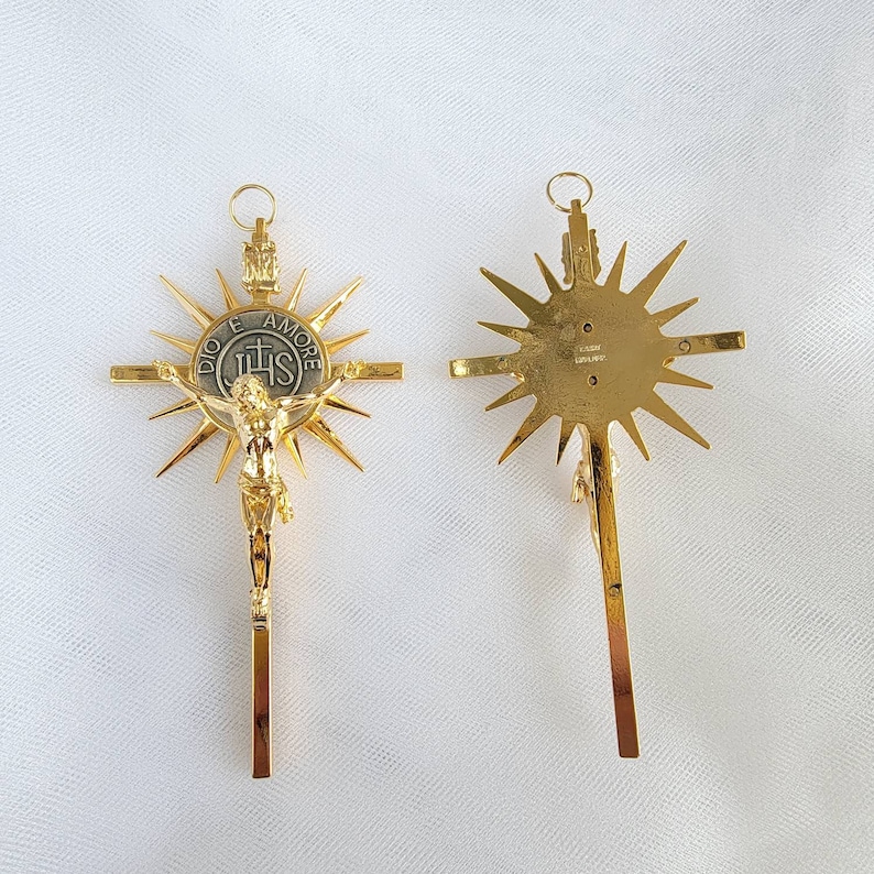 May include: Two gold-colored crucifixes. One shows a detailed depiction of Jesus Christ on the cross, with a sunburst design behind the cross. The other shows the back of the cross with a sunburst design. The crosses have a loop at the top for hanging.