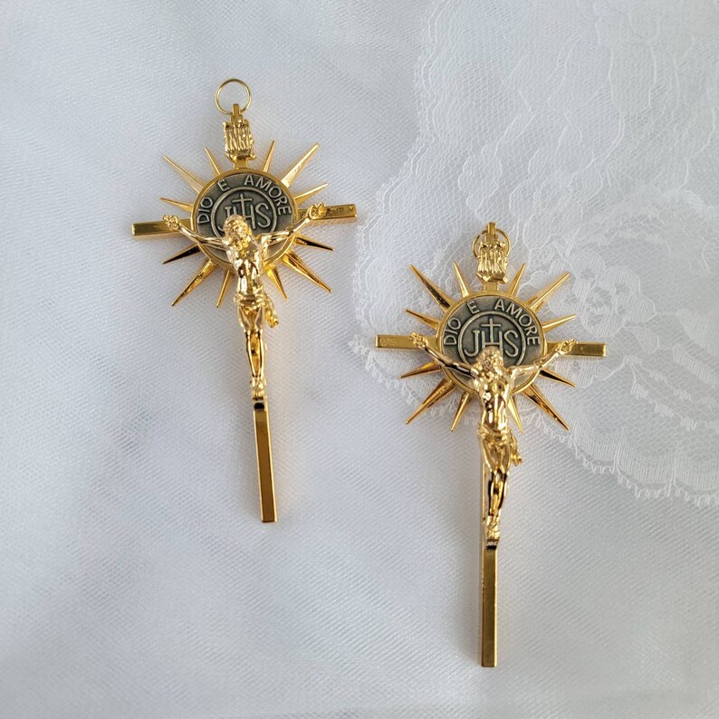 May include: Two gold-colored crucifix pendants. Each features a detailed depiction of Jesus Christ on the cross, with a sunburst design behind the cross. The center of the cross has the text "DIO E AMORE" and "IHS".