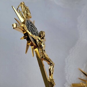 May include: A gold-colored crucifix with a detailed figure of Jesus Christ. The cross features a sunburst design and a circular emblem with text. The figure is highly detailed, with a polished finish. The overall design is ornate and religious.